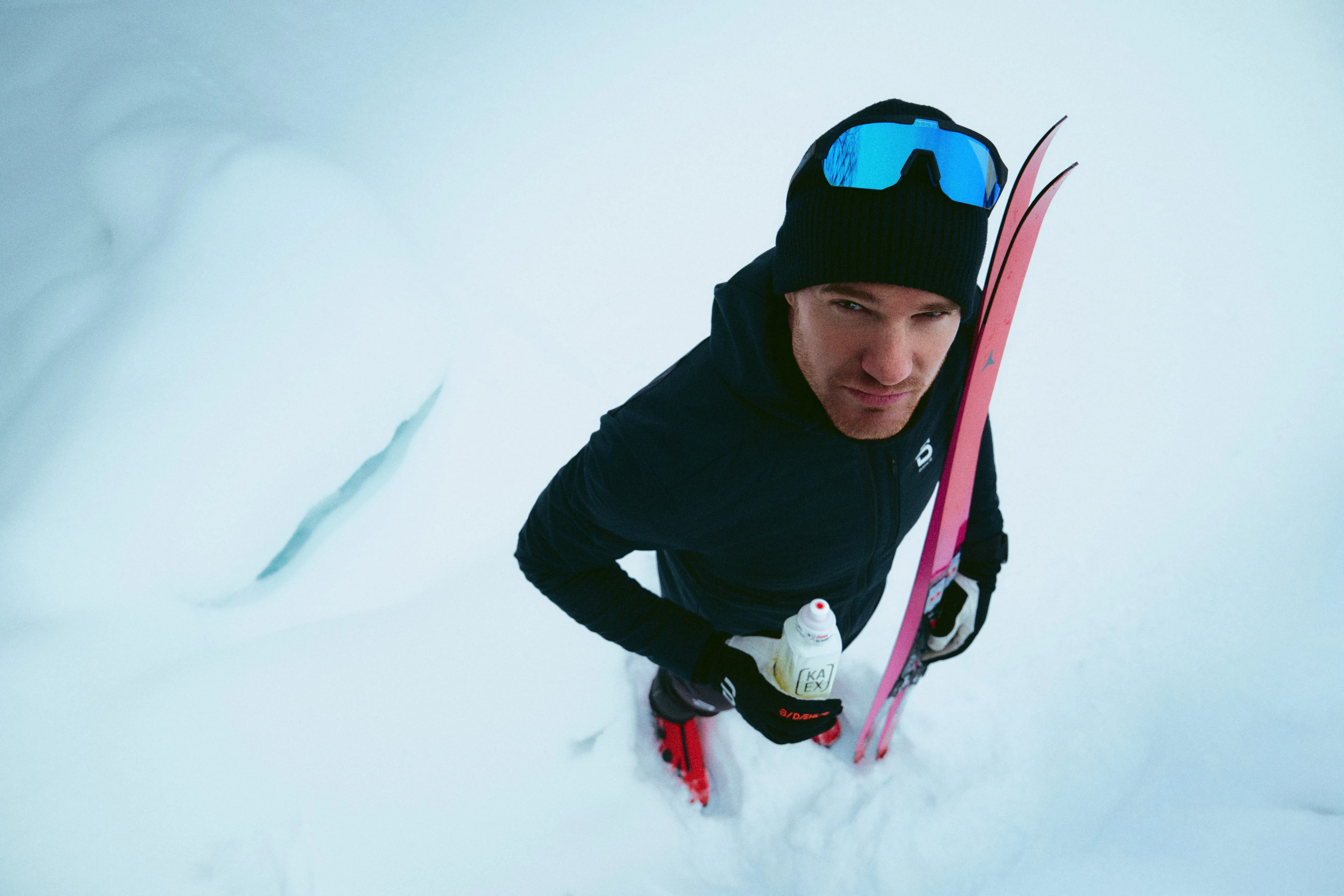 A top-down portrait of olympic champion Dario Cologna cross-country skier standing in deep snow, holding pink racing skis. He is wearing professional winter gear, including a black beanie and blue mirrored performance sunglasses.