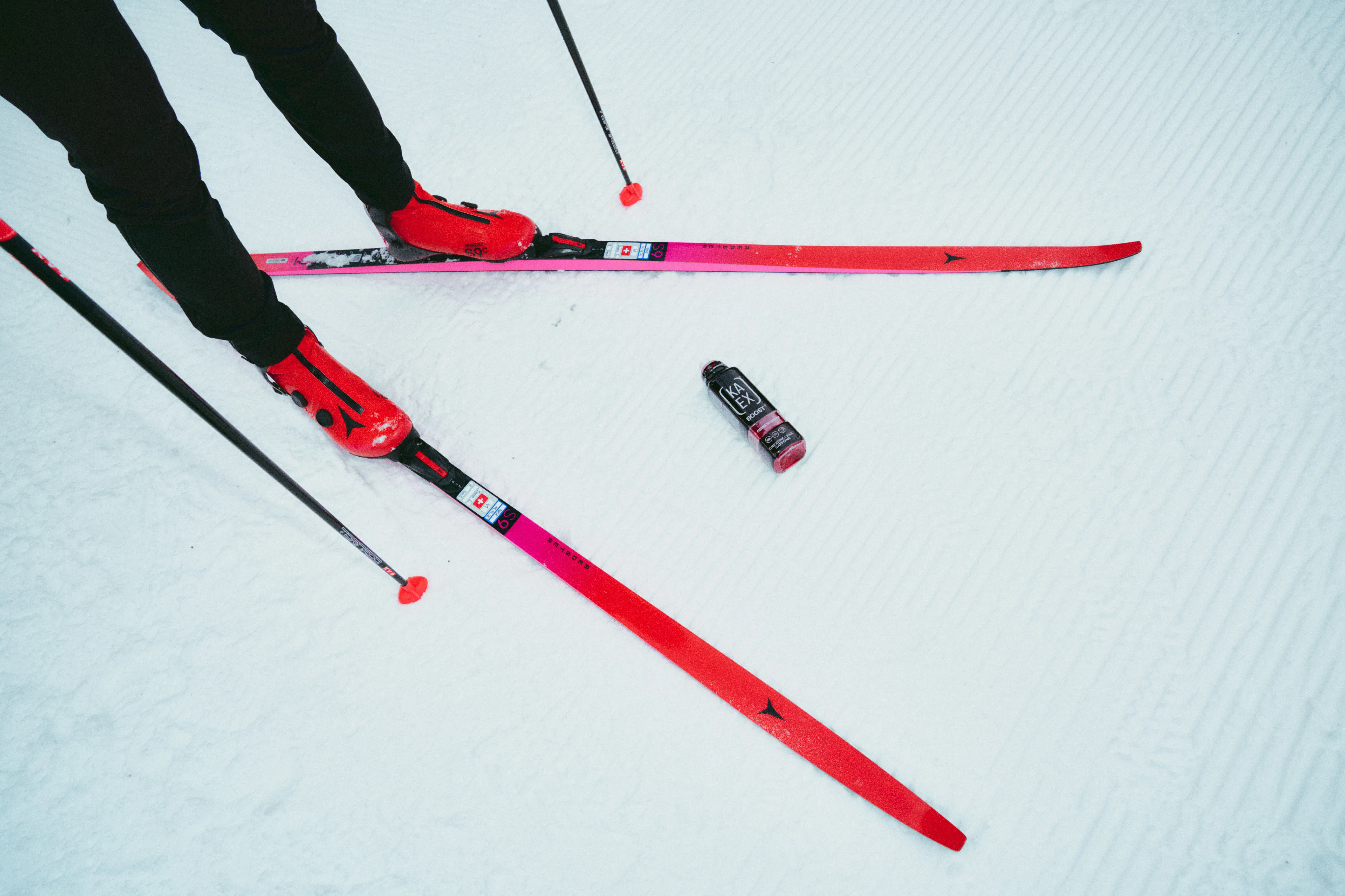 An action-oriented shot of a skier's legs and feet in red Atomic ski boots on a groomed snowy trail. A bottle of KAEX BOOST+ sits on the snow between vibrant pink skis, illustrating the integration of hydration and nutrition into winter training.