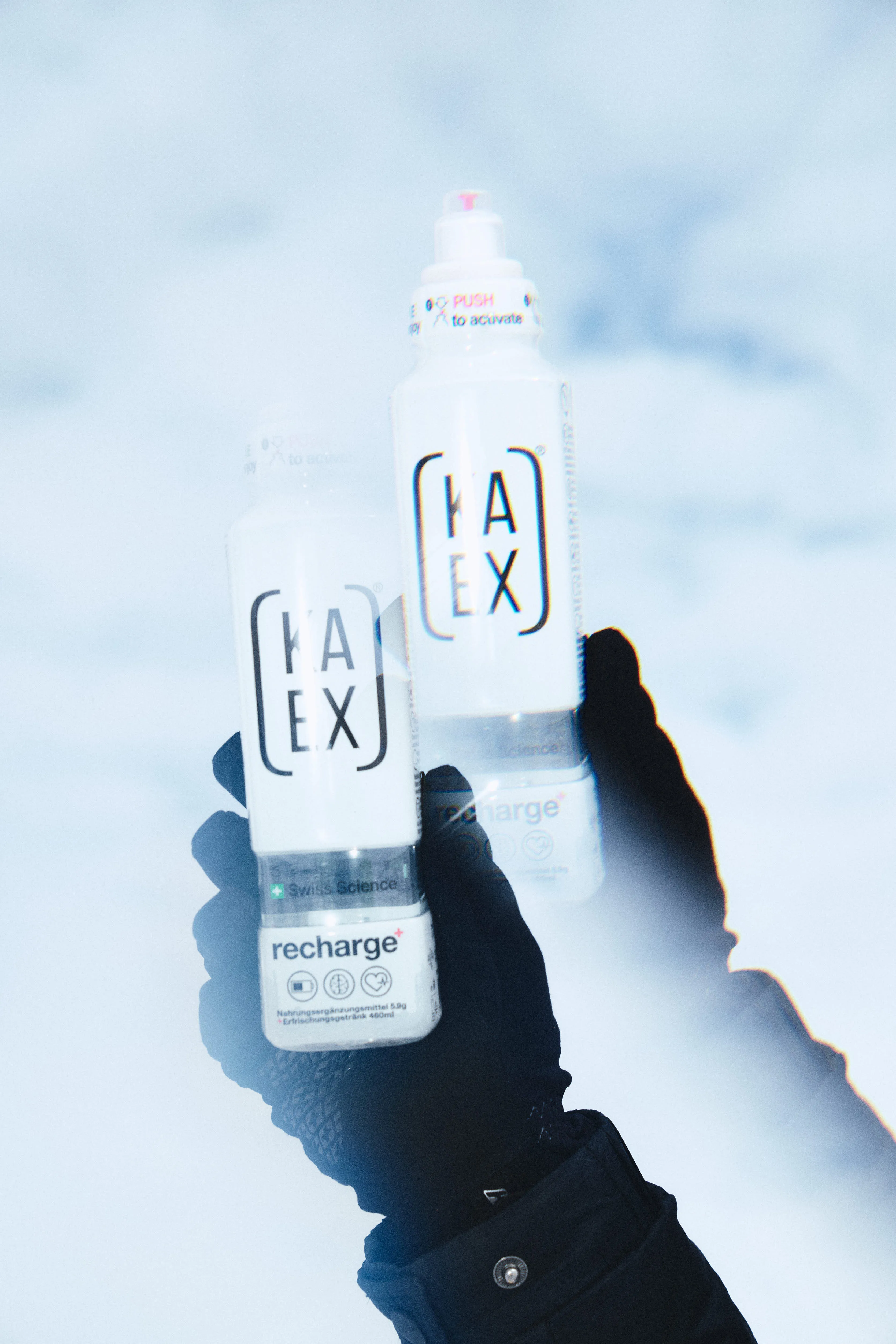 Hand holding two Kaex Recharge drink bottles in a snowy outdoor setting.