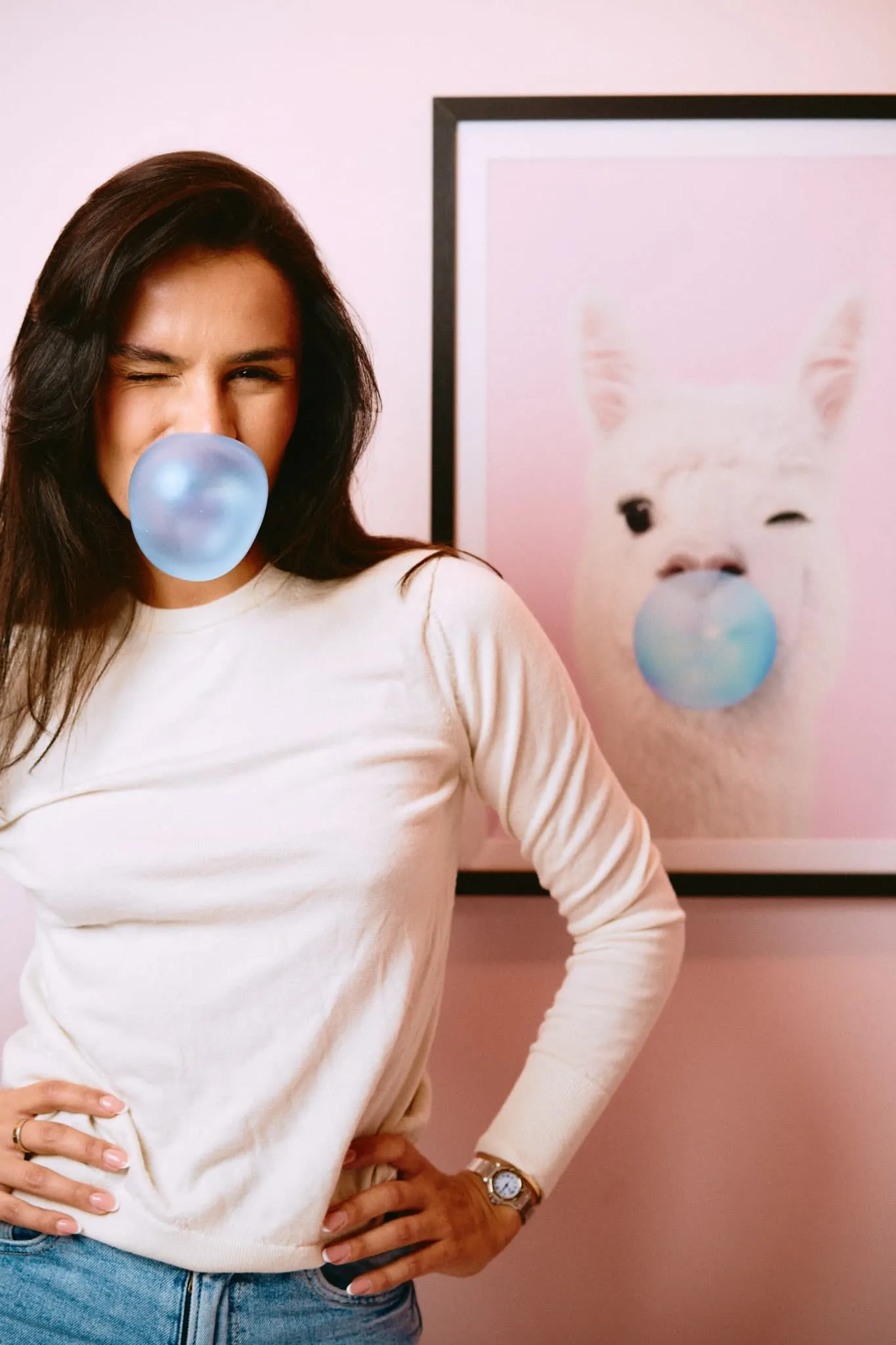A playful portrait of a woman blowing blue bubblegum in front of a matching framed photo of a llama doing the same — a lighthearted visual echo with personality and humor.