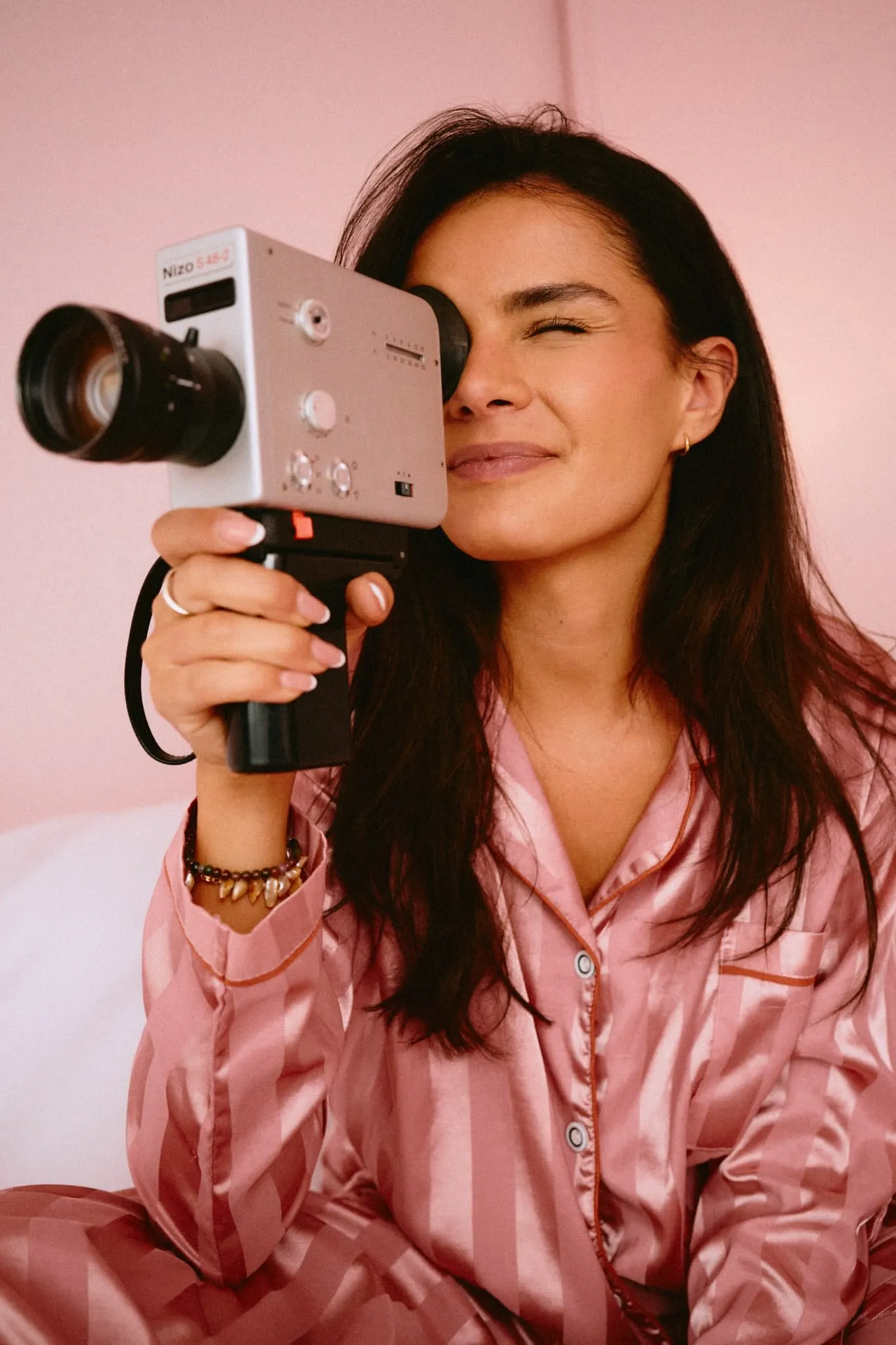A woman in striped pink satin pajamas smiles while filming with a vintage camera, capturing the joy of creativity in a cozy, pastel-toned setting.
